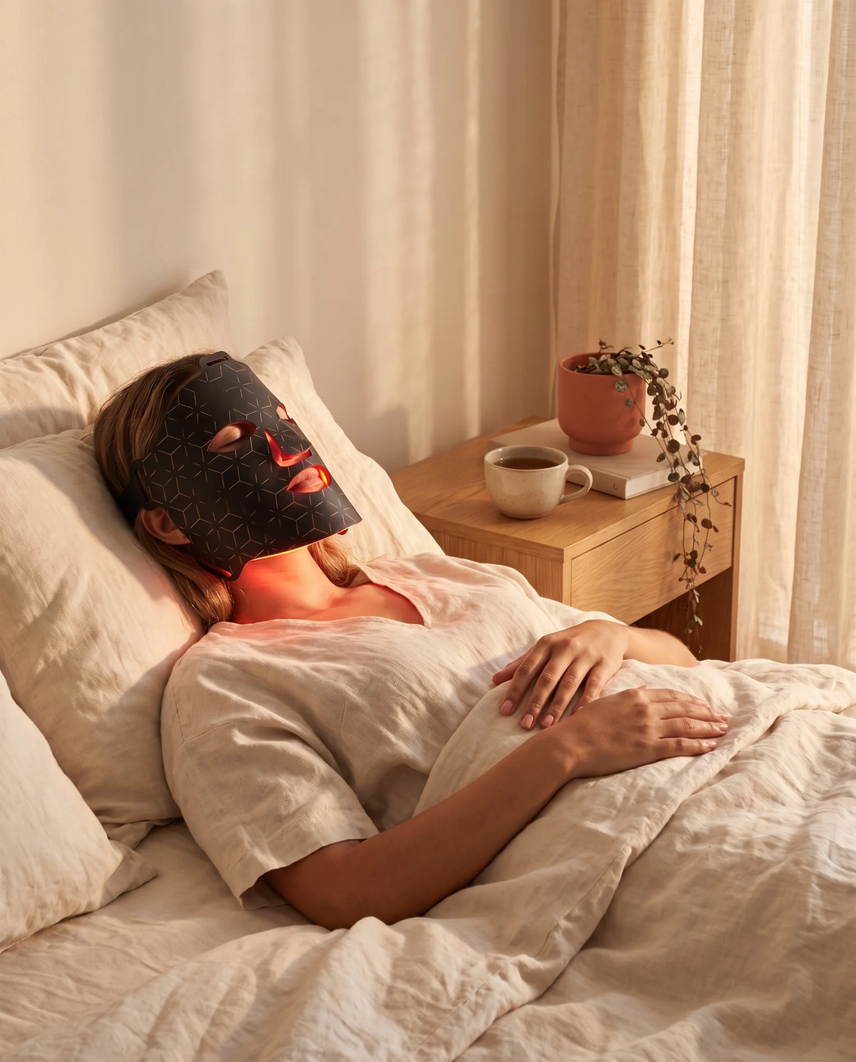 Woman resting in bed wearing the Clinirae Light Mask, glowing warm red